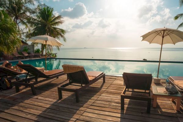 A serene poolside scene with wooden loungers, umbrellas, palm trees, and a view of a tranquil sea under a partly cloudy sky.