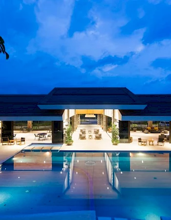 A luxurious building with a large outdoor swimming pool, surrounded by evening lighting under a partly cloudy sky.