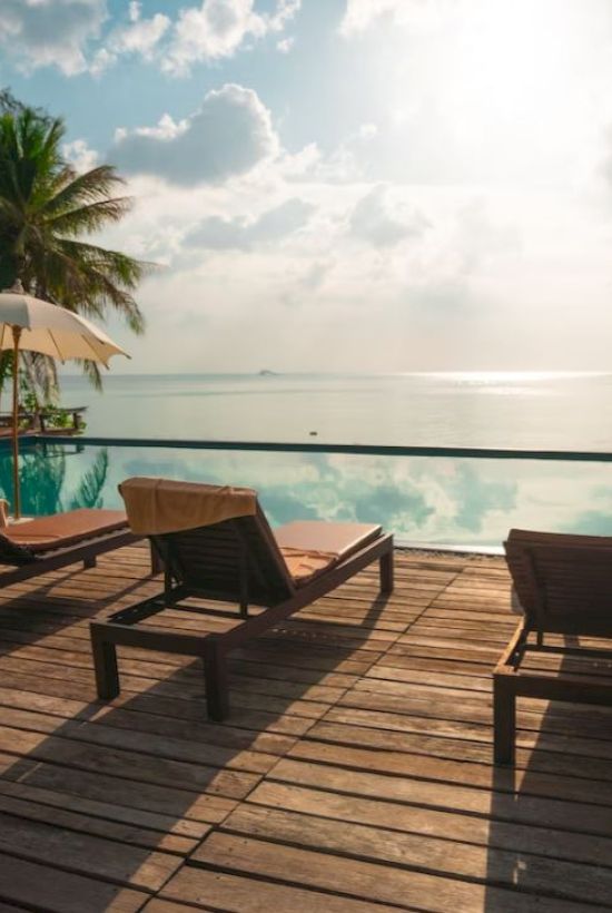 A tranquil poolside scene with lounge chairs, umbrellas, and a view of the ocean framed by palm trees under a sunny sky.