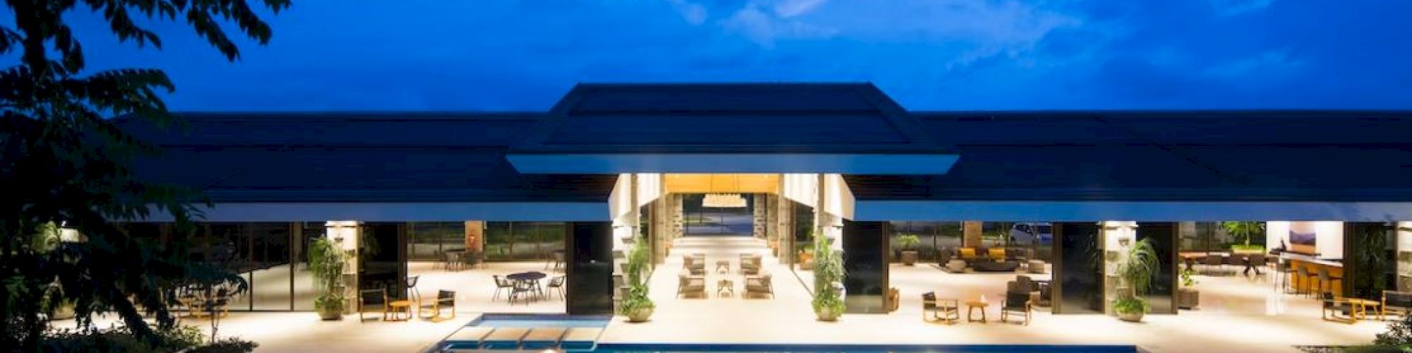 A modern building with large glass windows and an illuminated swimming pool at dusk, surrounded by outdoor seating.