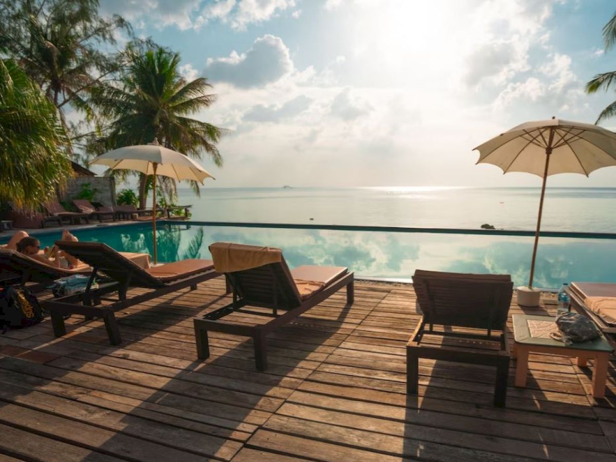 A serene beach scene with lounge chairs, umbrellas, a pool, palm trees, and a view of the ocean against a bright sky.