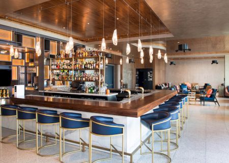 A modern bar with blue stools, elegant lighting, and a well-stocked counter, surrounded by upscale seating in a spacious setting.