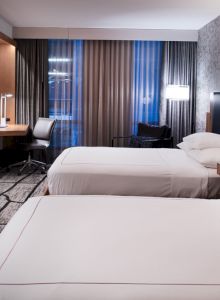The image shows a modern hotel room with two beds, a TV, a desk, and a patterned carpet, featuring warm lighting and stylish decor.