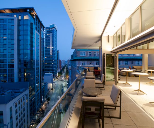 Modern rooftop terrace at dusk with seating, overlooking a cityscape of tall buildings and streets below, illuminated by city lights.