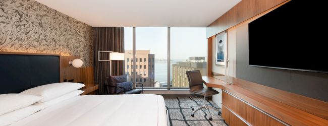 A modern hotel room features a large bed, wall-mounted TV, desk, chair, and a window with a cityscape view overlooking a body of water.