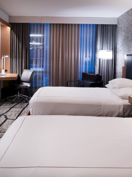 The image shows a modern hotel room with two beds, a desk, chair, TV, and decorative lighting. The furnishings are sleek and contemporary.