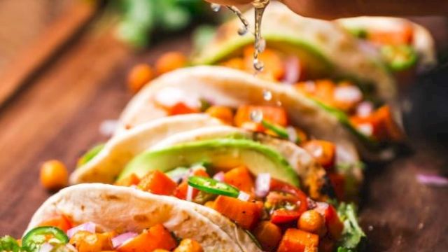 A hand squeezes lime over tacos filled with chickpeas, avocado, and vegetables on a wooden board, garnished with cilantro.