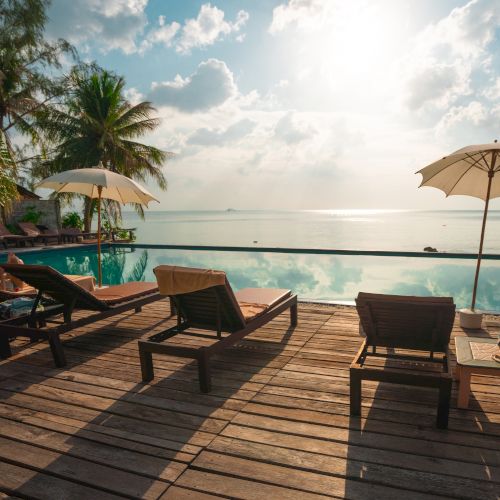 Beachside scene with lounge chairs and umbrellas on a wooden deck by the sea, under a partly cloudy sky with palm trees.