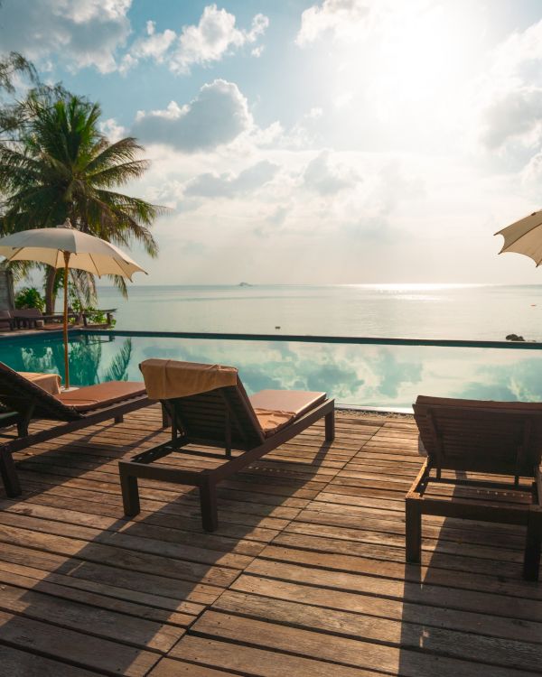 Beachside scene with lounge chairs and umbrellas on a wooden deck by the sea, under a partly cloudy sky with palm trees.