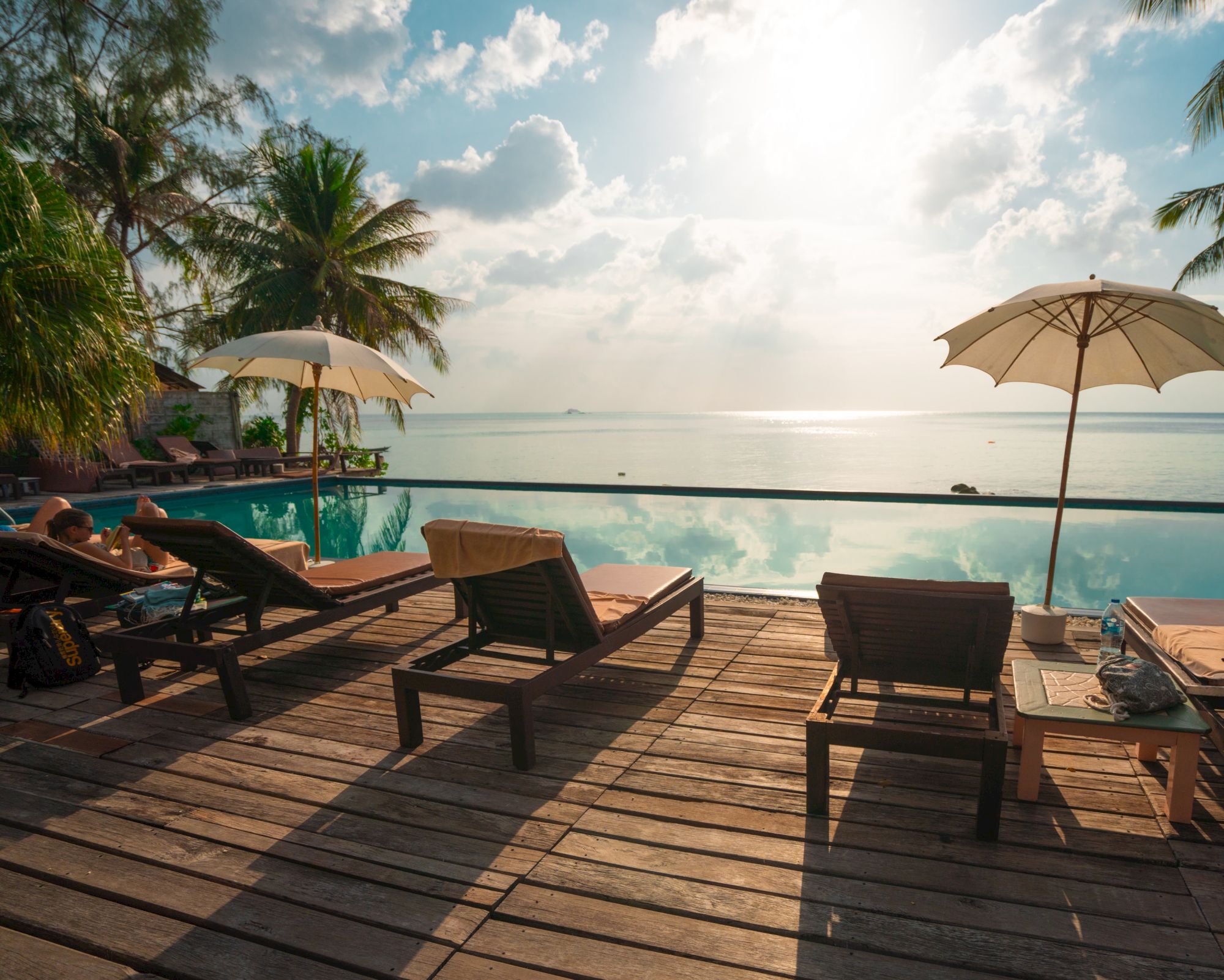 A serene beachfront scene with lounge chairs, umbrellas, and palm trees by an infinity pool overlooking a calm ocean and a bright sky.