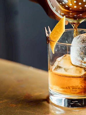 A cocktail being poured into a glass with ice and a lemon twist on the rim.