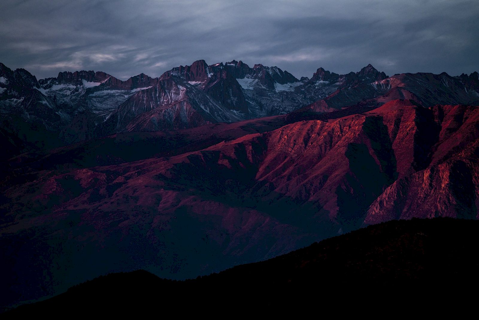 A mountain range bathed in pink and purple hues under a cloudy sky during sunset, showcasing dramatic light and shadow contrasts.