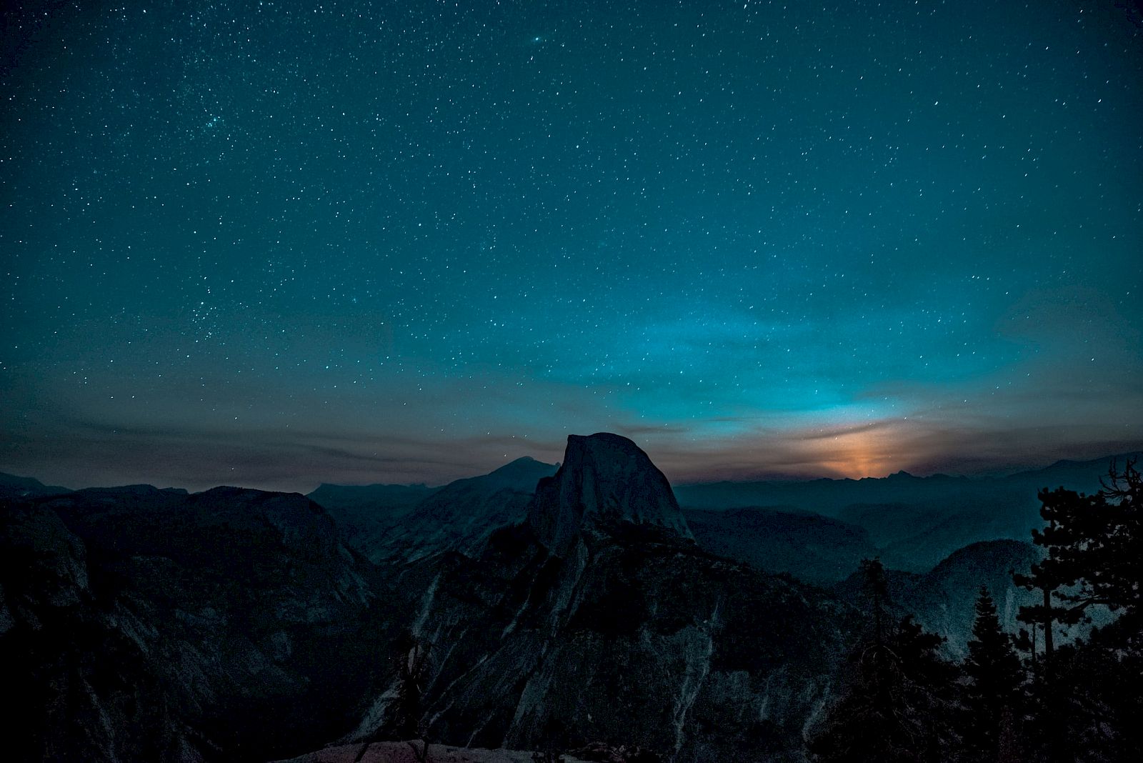 A starry night sky over rugged mountains, with a faint glow on the horizon, creating a serene and captivating landscape.