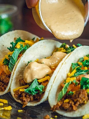 Three tacos filled with ground meat, cheese, greens, and tomatoes, being drizzled with creamy sauce on a plate.