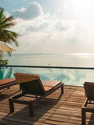 Poolside with deck chairs, umbrellas, and a view of palm trees and the sea under a cloudy sky. Relaxing tropical getaway atmosphere.