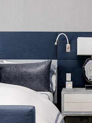 A stylish bedroom with a dark blue headboard, white bedding, pillows, a bedside table, modern lamp, and a wall-mounted reading light.