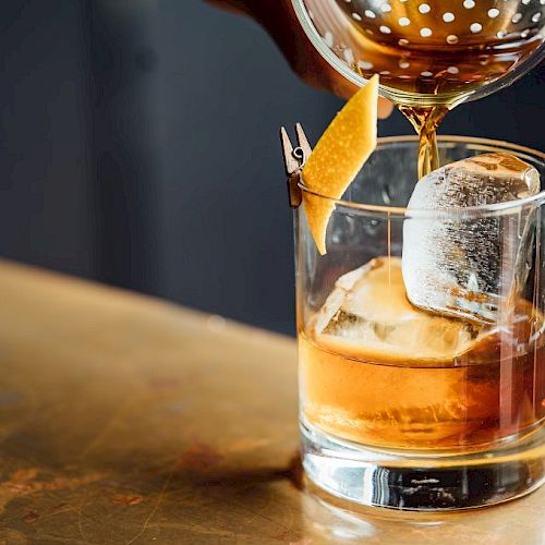 A cocktail being poured into a glass with a large ice cube and a citrus twist garnish, placed on a wooden surface.