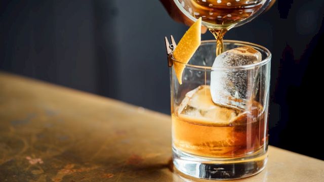 A cocktail being poured into a glass with a large ice cube and a citrus twist garnish, placed on a wooden surface.