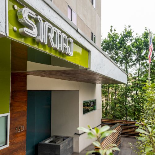 The image shows the entrance of a building with the sign "SIRTAJ" and surrounded by greenery.