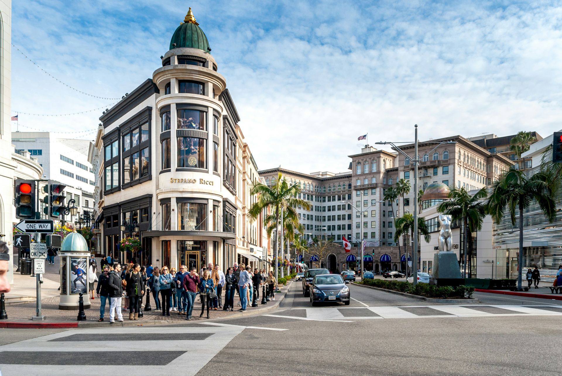 A bustling city intersection with a historic building, palm trees, and people walking around. Cars are waiting at the traffic light.