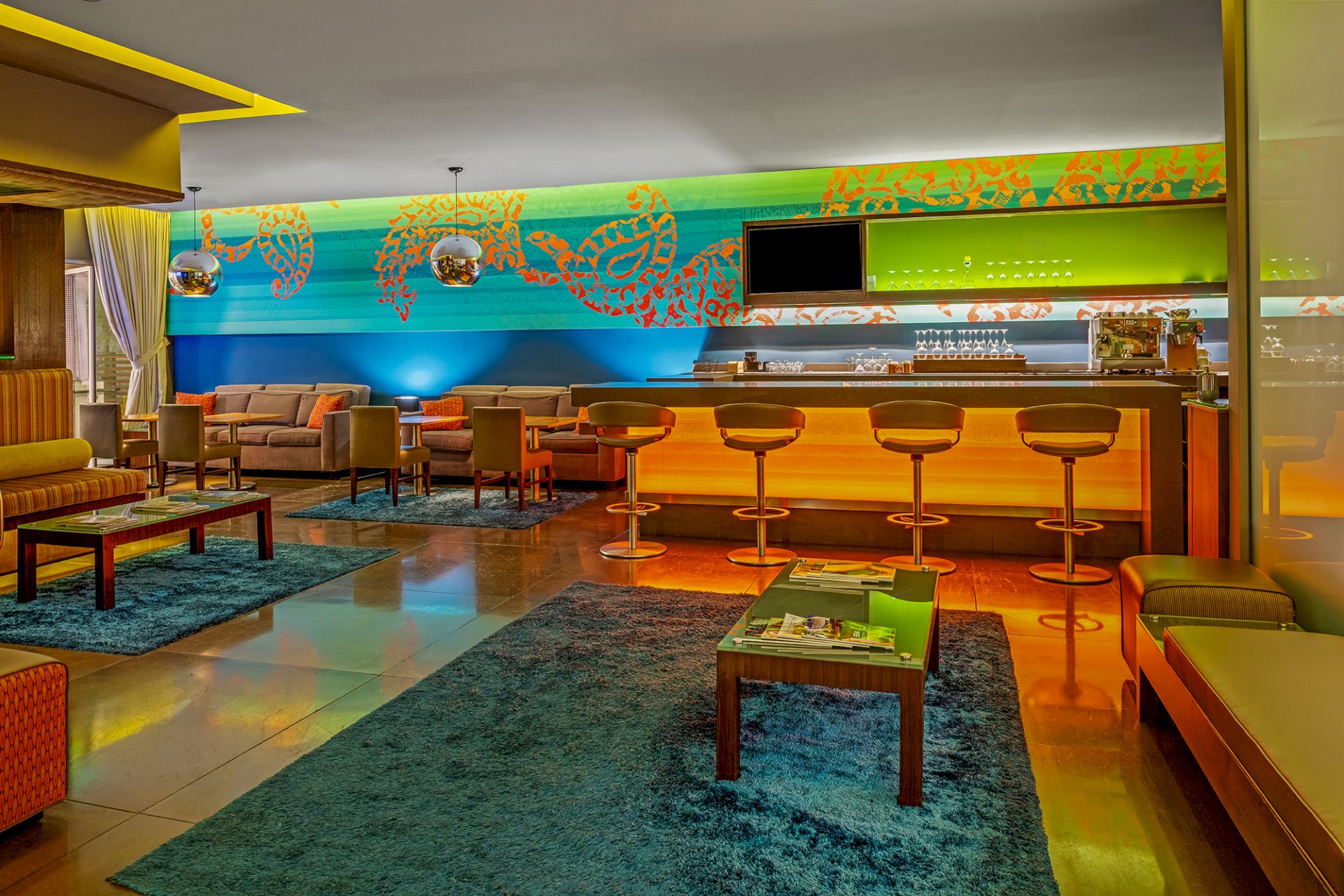 A modern lounge with a bar, colorful lighting, bar stools, couches, a television, and vibrant wall art.
