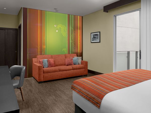 A modern hotel room features a bed with an orange-striped cover, an orange sofa, and a desk with a chair, with a balcony access.