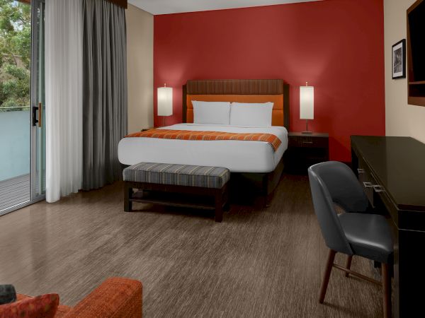 A modern hotel room with a bed, desk, chair, and a balcony. The room has red and cream walls, wooden flooring, and a large window.