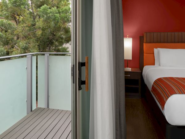 The image shows an open balcony door leading to a wooden deck, with a modern bedroom featuring a bed and orange accents visible inside.