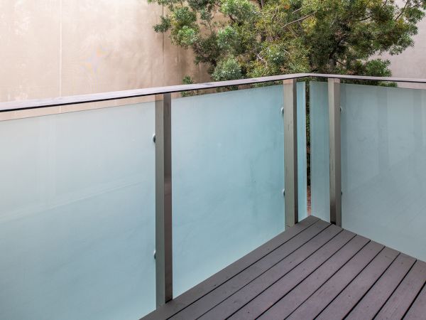 A small balcony with frosted glass panels and metal railings, featuring wooden flooring and a tree visible in the background.