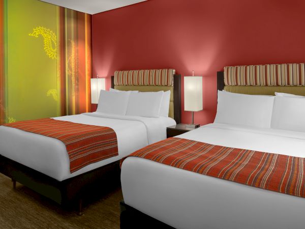 The image shows a hotel room with two neatly made double beds, red-striped bed runners, modern lamps, and colorful wall decor.
