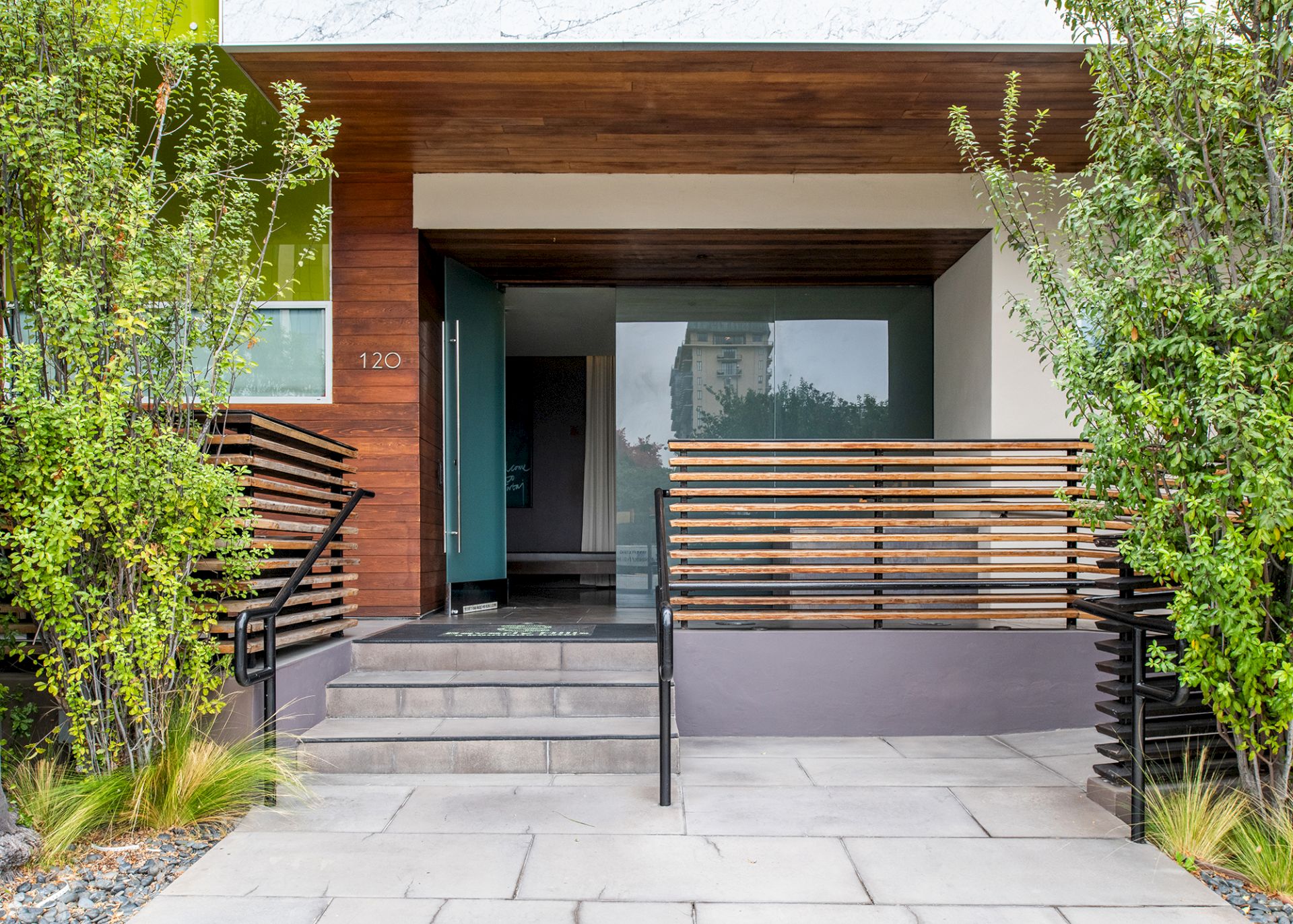 The image shows a modern building entrance with a wooden facade, glass door, steps, and greenery on both sides, creating a welcoming look.