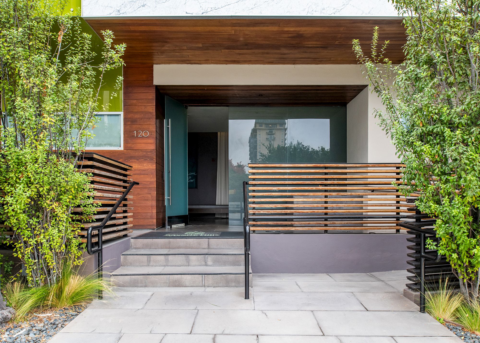The image shows a modern building entrance with a wooden facade, glass door, steps, and greenery on both sides, creating a welcoming look.