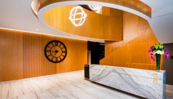 A modern lobby with wooden paneling, a marble desk, a large clock, and stylish lighting. A vase with flowers is on the desk.