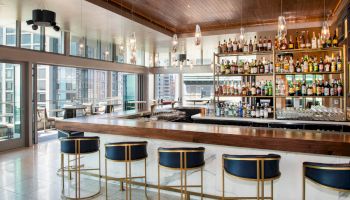 A modern bar with a long counter, high chairs, and shelves stocked with various bottles against large windows in an urban setting.