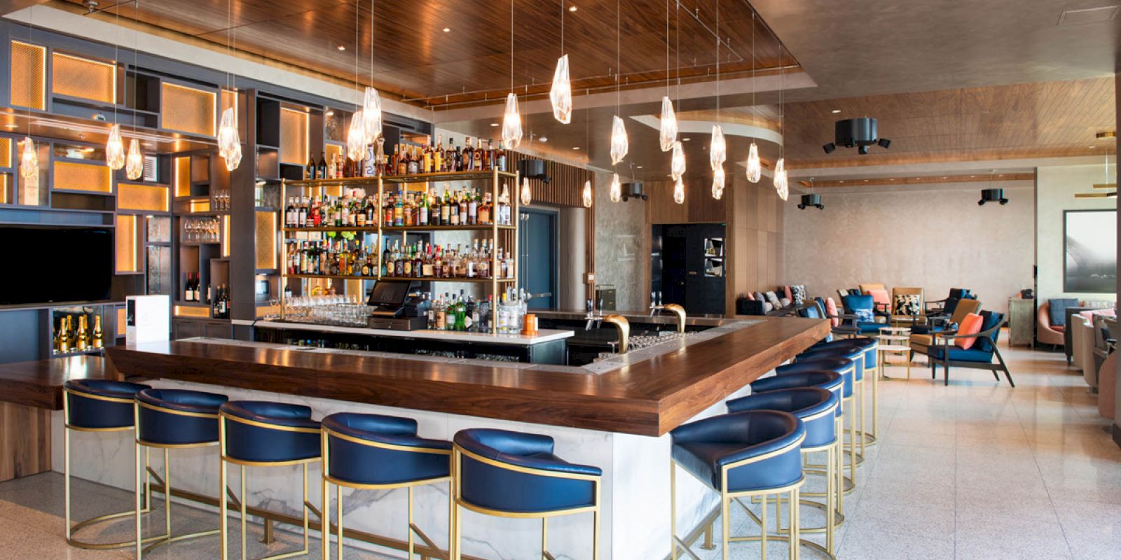 A modern bar with a U-shaped counter, blue stools, pendant lights, and a well-stocked bar display. Lounge seating is visible in the background.