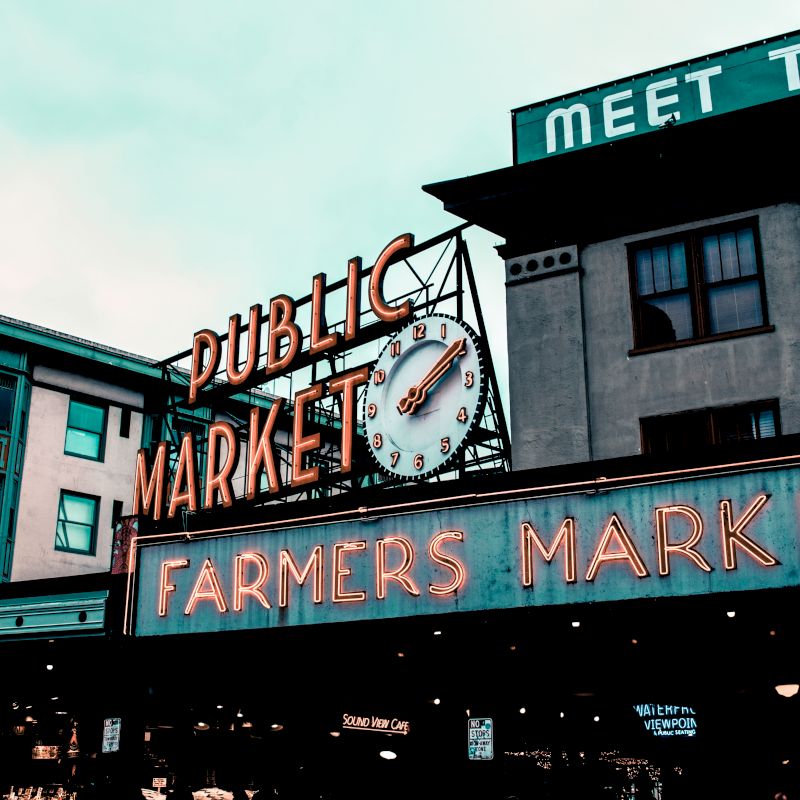 The image features a public market sign with a clock and neon-lit "Farmers Market," set against an urban backdrop, probably in Seattle.