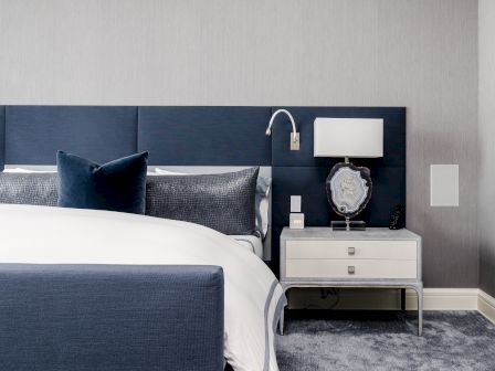 A modern bedroom features a navy bed, white bedding, a sleek nightstand with a lamp, and a decorative item in a neutral-toned setting.