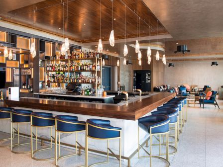 A modern bar with blue stools, a well-stocked liquor shelf, and pendant lights, set in a cozy lounge area with seating.