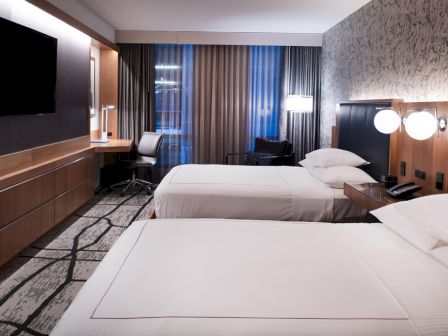 The image shows a modern hotel room with two beds, a wall-mounted TV, a work desk, and lamps, decorated in neutral tones and patterns.