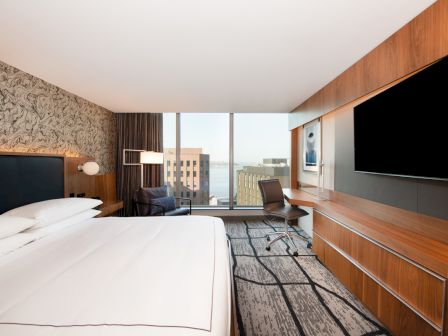 A modern hotel room with a large bed, desk, chair, and TV. There’s a view of buildings and water through a window.
