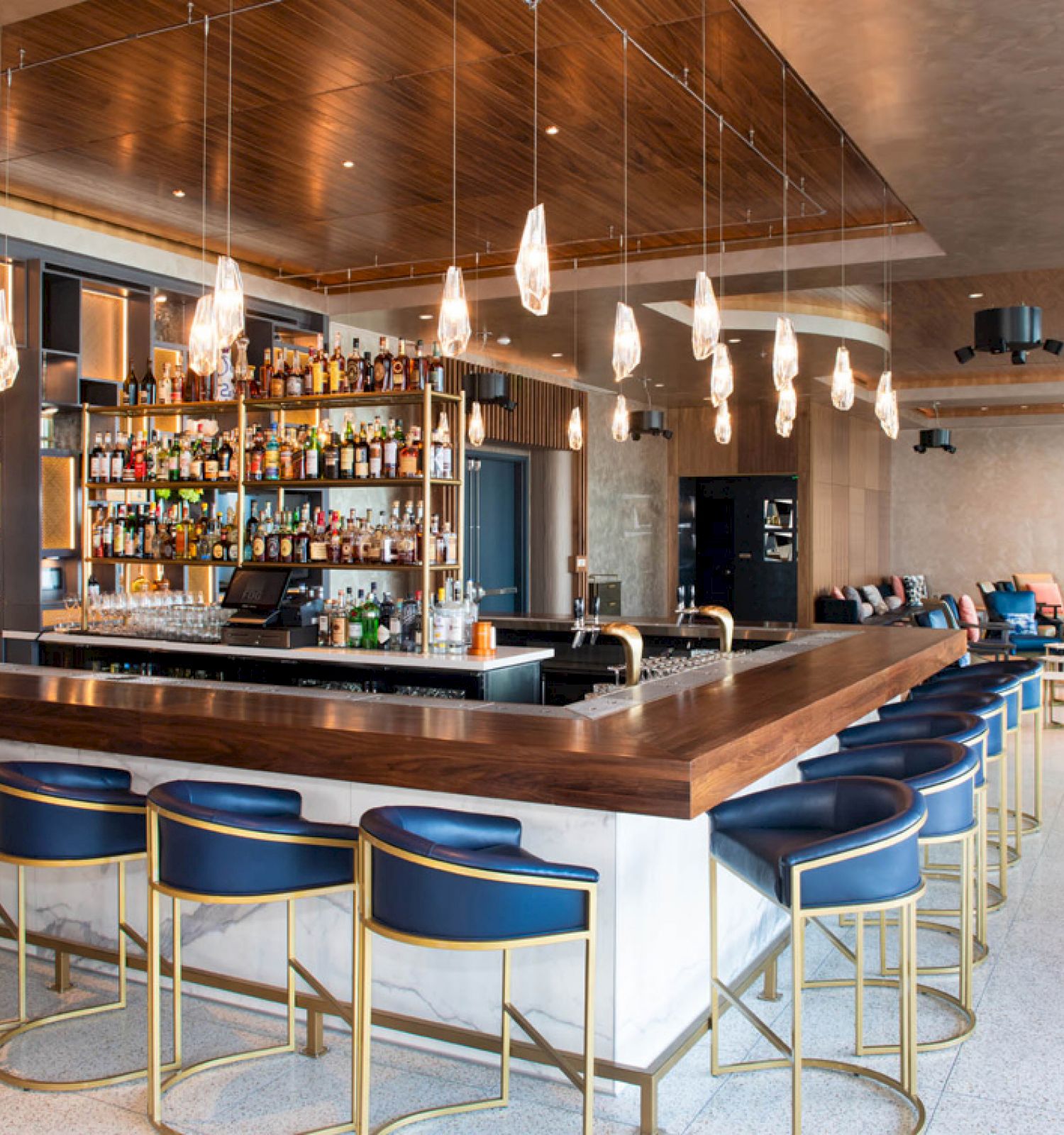 This image shows a modern bar with blue stools, a wooden counter, and a variety of liquor bottles on shelves, complemented by pendant lights.