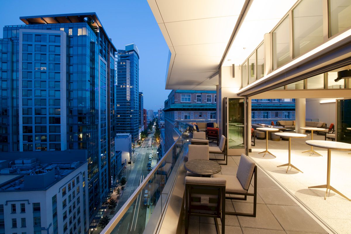 The image shows a modern cityscape with high-rise buildings and a luxurious balcony with seating and tables in an urban setting at dusk.