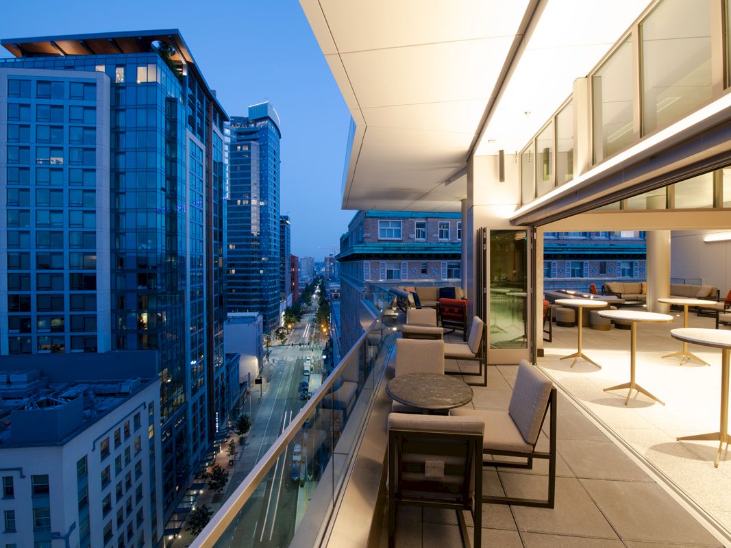 The image shows a modern cityscape with high-rise buildings and a luxurious balcony with seating and tables in an urban setting at dusk.