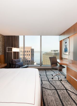The image shows a modern hotel room with a large bed, a flat-screen TV, a work desk, and a city view through the windows.