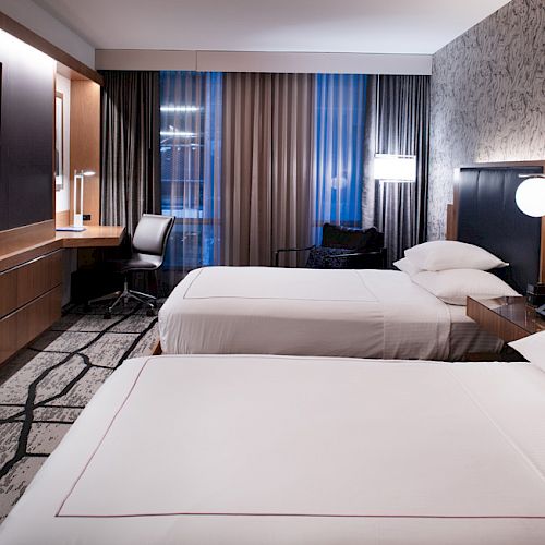 A modern hotel room with two beds, a wall-mounted TV, a desk, a chair, and stylish lighting. A patterned carpet and decorative wallpaper complete the look.