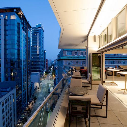 A modern balcony overlooking a cityscape at dusk, with outdoor seating and glass railings, facing illuminated buildings across the street.