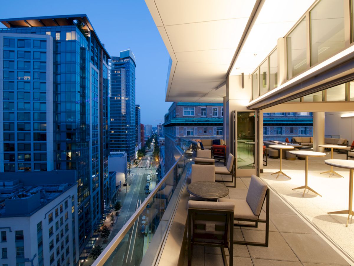 Cityscape at dusk with modern high-rise buildings, a balcony with seating and tables, and a view of the street below.