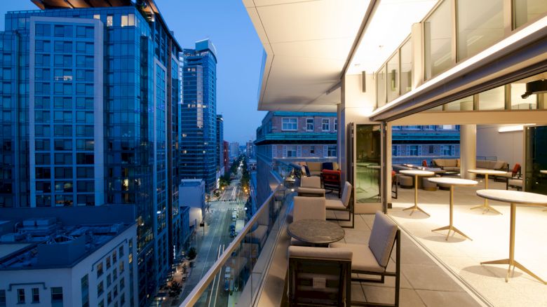 Cityscape at dusk with modern high-rise buildings, a balcony with seating and tables, and a view of the street below.