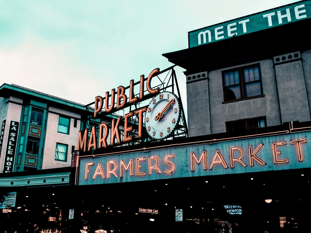 The image shows a neon sign for a public market and farmers market, with a clock, on an urban building's facade.