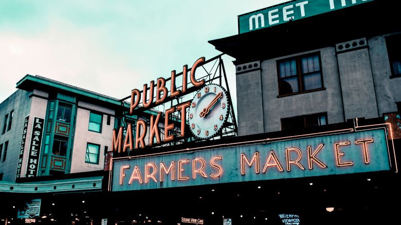 The image shows a neon sign for a public market and farmers market, with a clock, on an urban building's facade.